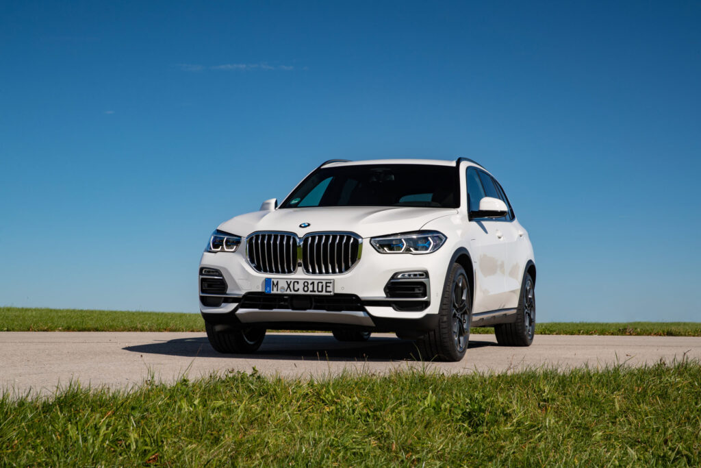 Best Luxury Hybrid SUV pictured as a white BMW X5 xDrive45e plug-in hybrid SUV parked on a quiet road with green grass and a clear blue sky.
