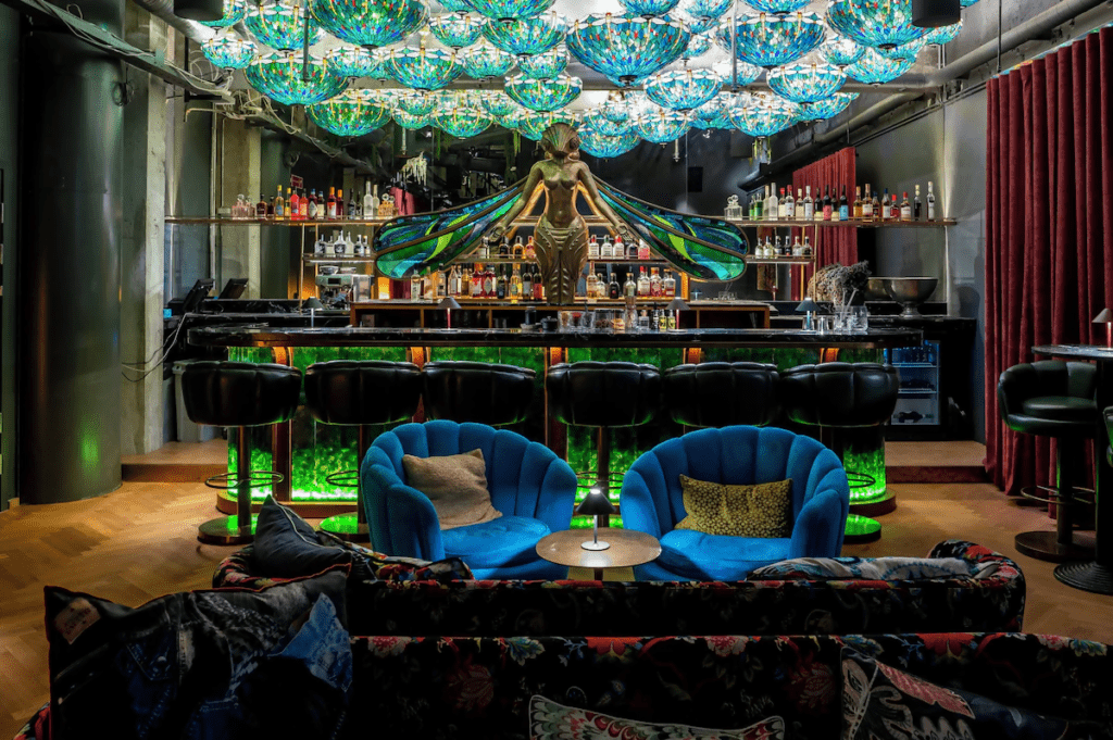 Design and atmosphere inside a bohemian luxury hotel bar with colorful mosaic pendant lights, a sculptural winged centerpiece behind the bar, green-lit bar stools, and plush blue velvet lounge chairs.