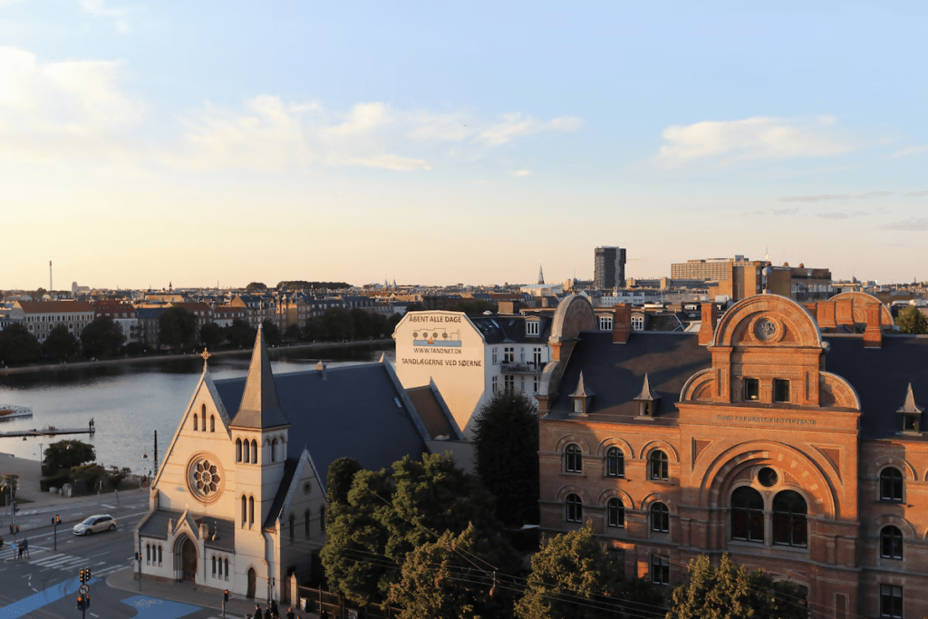Location and accessibility view of central Copenhagen skyline at sunset, overlooking a lake, historic church spire, and classic brick buildings near the city center.