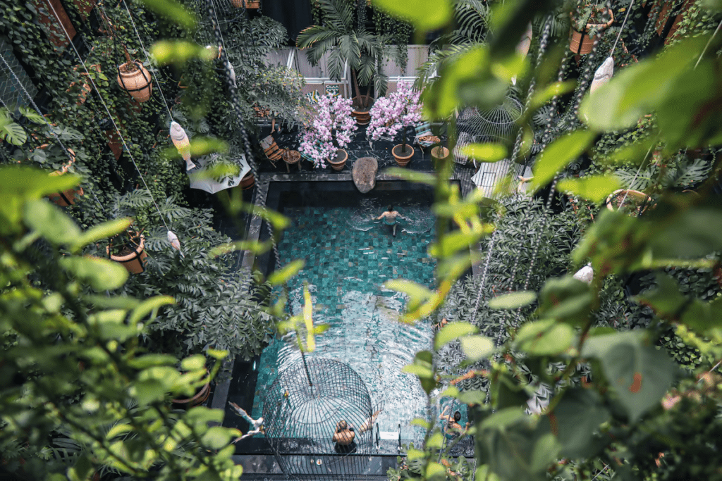 Manon Les Suites Hotel indoor courtyard pool seen from above, teal mosaic water surrounded by lush tropical greenery, hanging plants, and pink flowering trees, creating a serene urban oasis vibe.