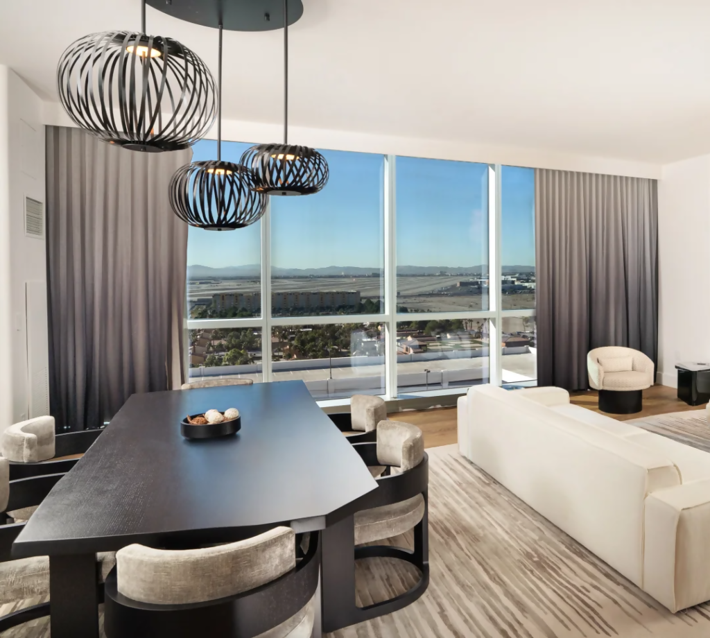 Modern hotel suite with black dining table, sculptural pendant lights, floor-to-ceiling windows, and a white sofa with a desert view.