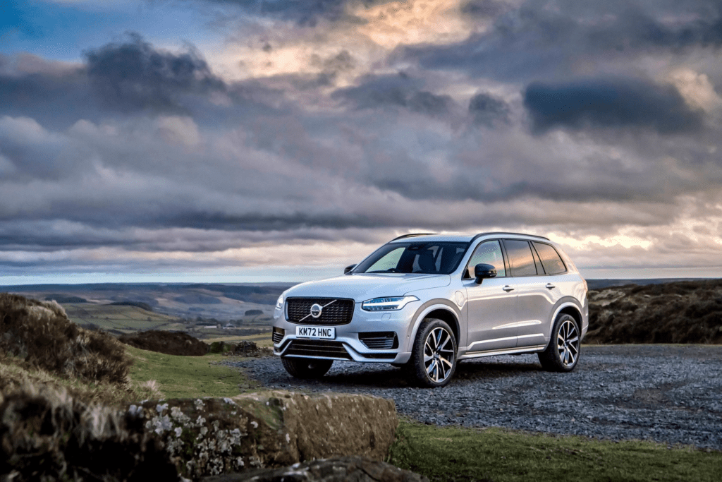 Best Luxury Hybrid SUV shown as a silver Volvo XC90 Recharge plug-in hybrid SUV parked on a scenic hillside road under dramatic cloudy skies.